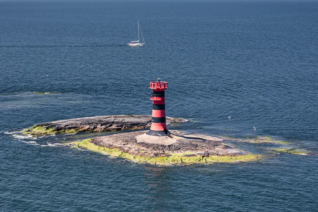 Leuchtturm auf einem kleinen Felsvorsprung im Wasser, umgeben von grünen Algen. Im Hintergrund segelt ein Boot.