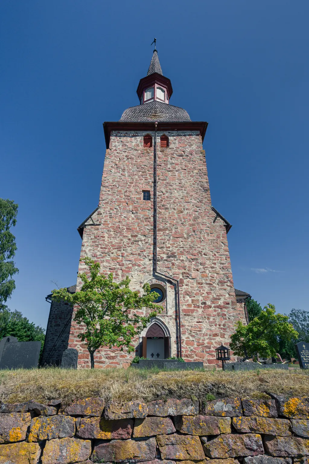 Steinerne Kirche mit hoher spitz zulaufender Turmhaube, umgeben von Bäumen und einer niedrigen Steinmauer. Klarer blauer Himmel.
