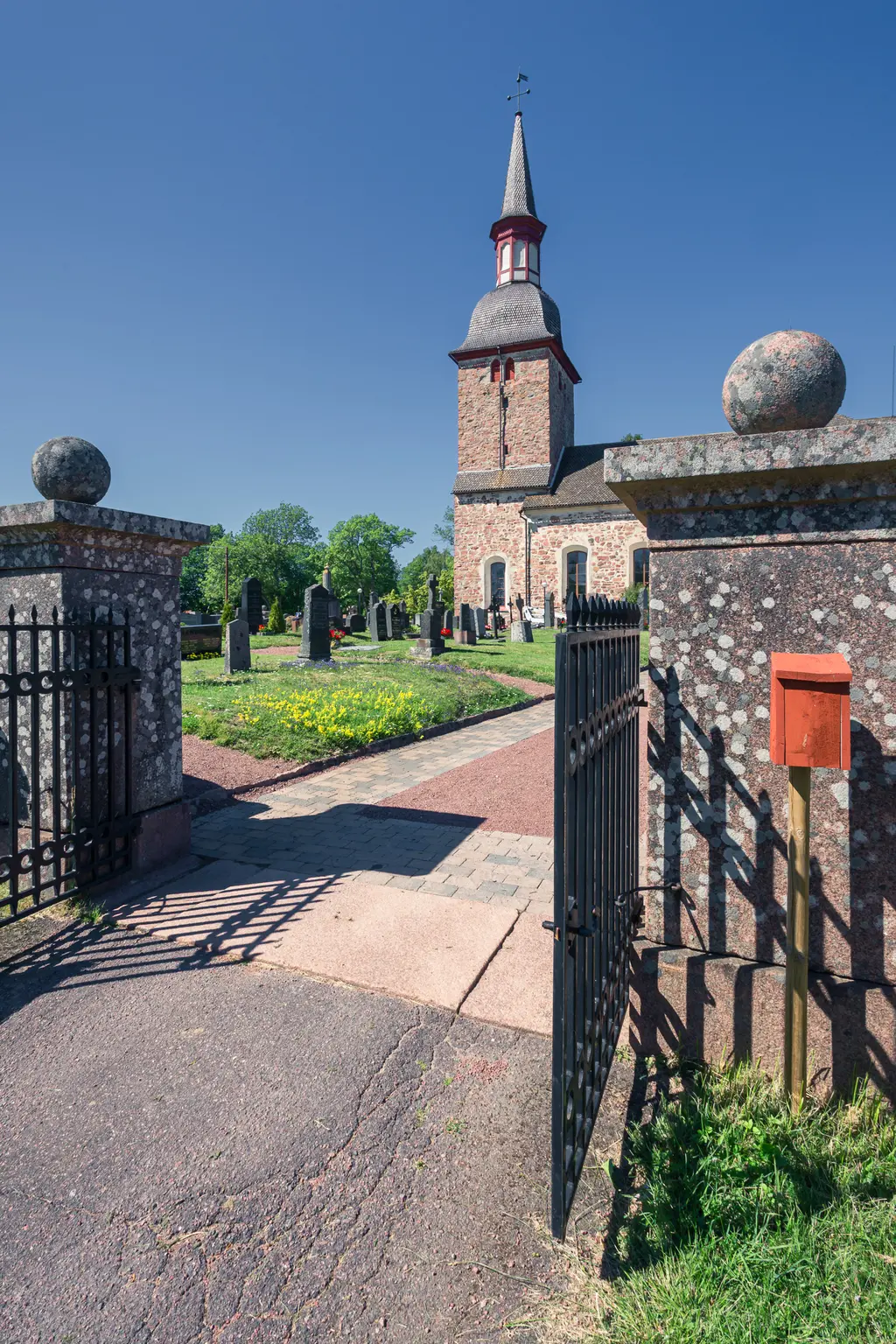 Historische Kirche mit spitzem Turm, umgeben von einem Friedhof. Ein gepflasterter Weg führt durch ein schmiedeeisernes Tor. Grüne Wiese und gelbe Blumen sind sichtbar.