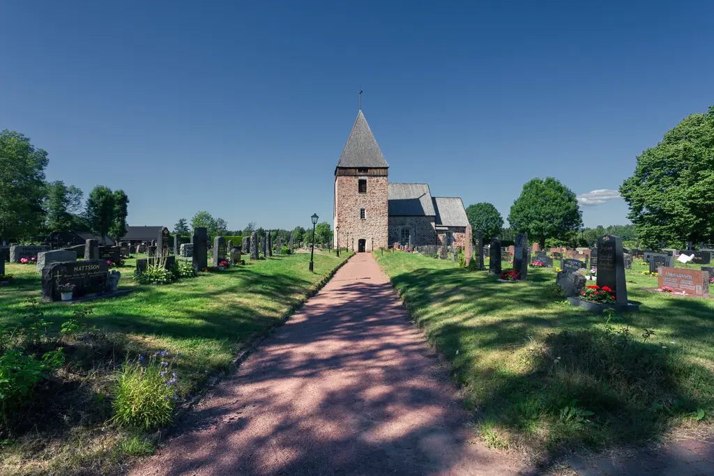 Historische Kirche mit rechteckigem Turm, umgeben von einem gepflegten Friedhof. Ein gepflasterter Weg führt zur Kirche, von Bäumen gesäumt.