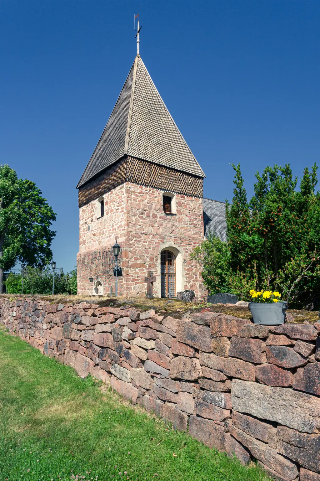 Historisches Gebäude mit schiefergedecktem, spitzen Turm und schmalen Fenstern, umgeben von einer niedrigen Steinmauer und grünem Gras.