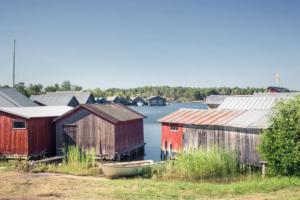Rote Holzschuppen am Wasser mit grünem Gras und einem kleinen Boot. Im Hintergrund sind weitere Gebäude und Bäume sichtbar.