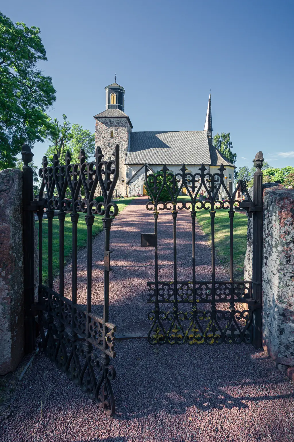 Historische Kirche hinter einem kunstvollen, offenen Eisengittertor. Der Weg zur Kirche ist mit kleinen Steinen bedeckt und von Bäumen umgeben.