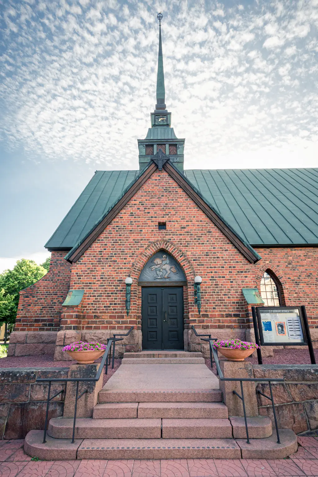 Ziegelsteinkirche mit spitzem Turm und breiten Treppenstufen, umgeben von Blumenbeeten. Die Fassade zeigt architektonische Details und Fenster.