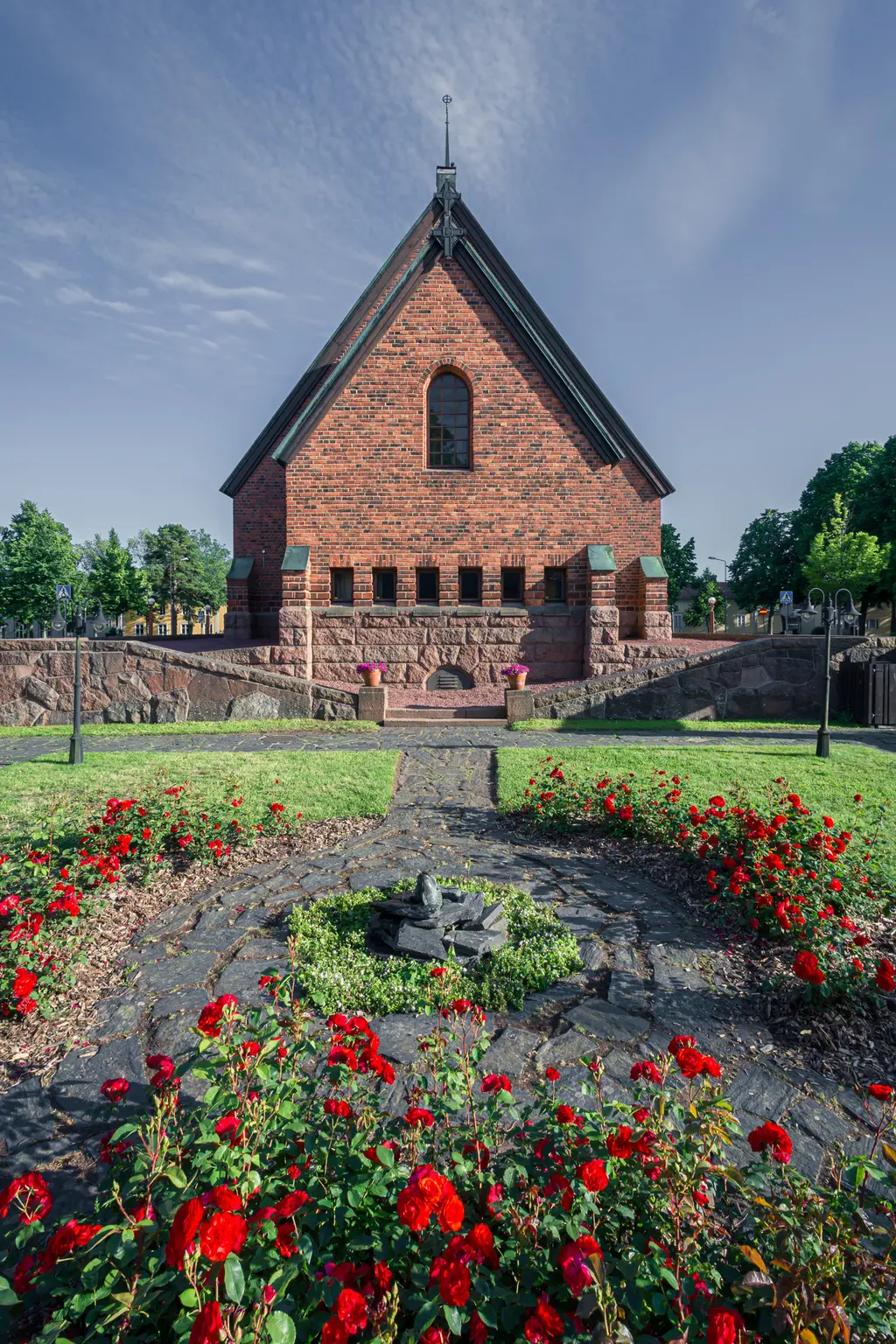 Backsteekirche aus roten Ziegeln, umgeben von bunten Blumenbeeten und Grasflächen. Im Vordergrund ein Steinmuster.