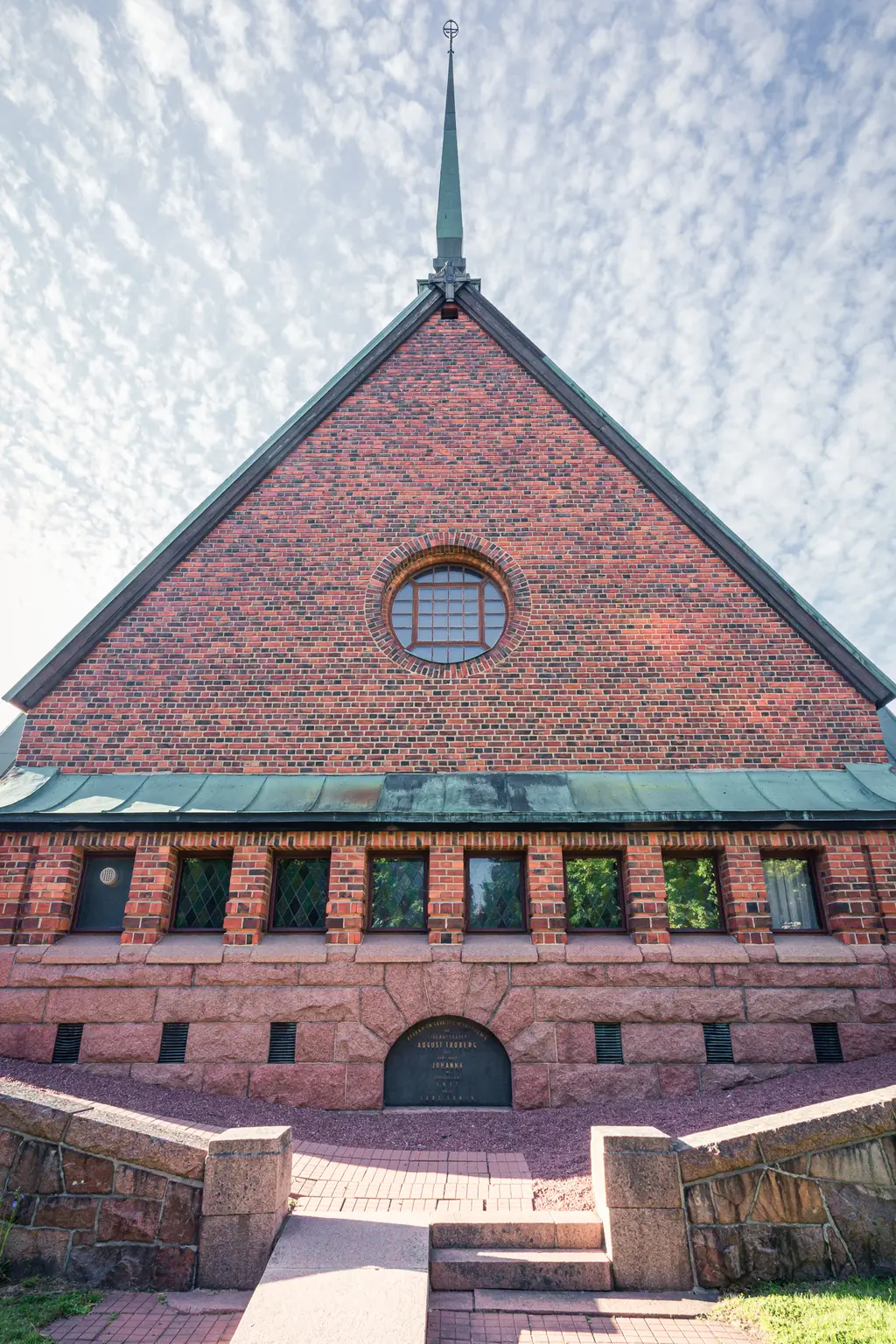 Rotziegelneues Gebäude mit spitzem Dach und einem hohen, grünen Dachreiter. Fenster in Bögen und rechteckiger Form, Steintreppe im Vordergrund.