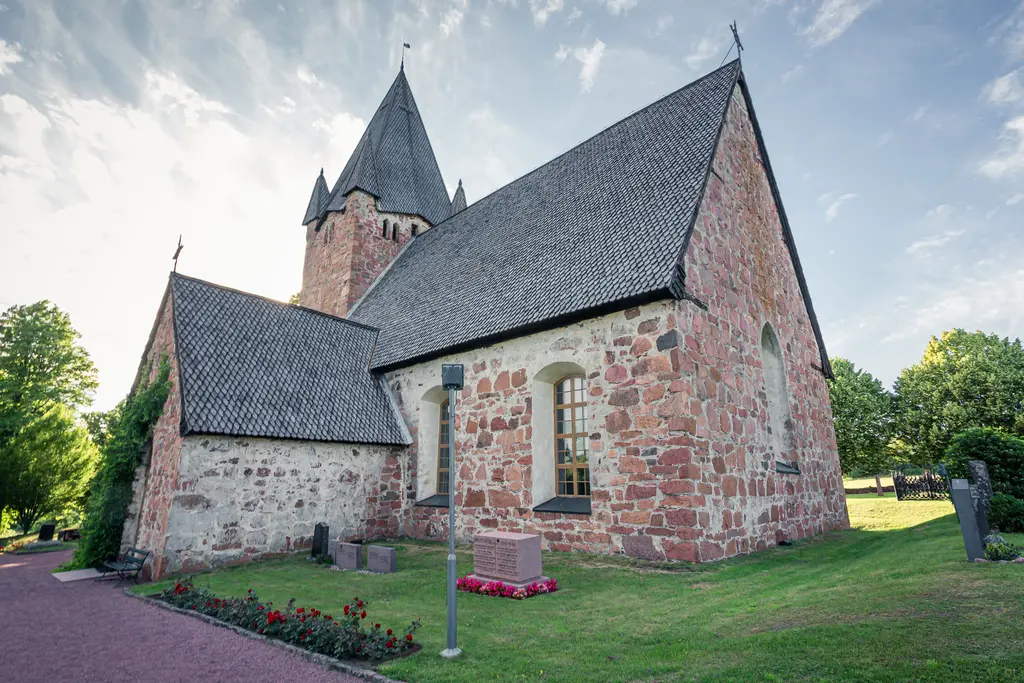 Historische Steinkirche mit einem hohen, spitzen Dach und einer umliegenden, grünen Wiese. Rosenbeet vor der Kirche.