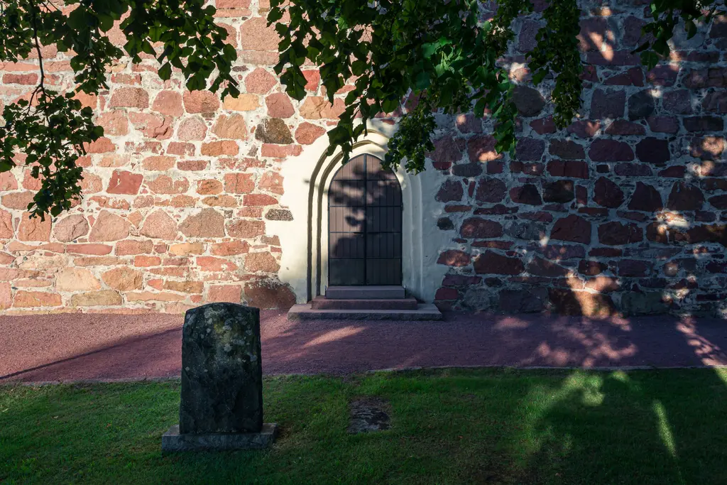 Steinwand einer historischen Kirche mit einer Holztür, daneben ein kleiner Grabstein. Schatten von Bäumen fallen auf den Boden.