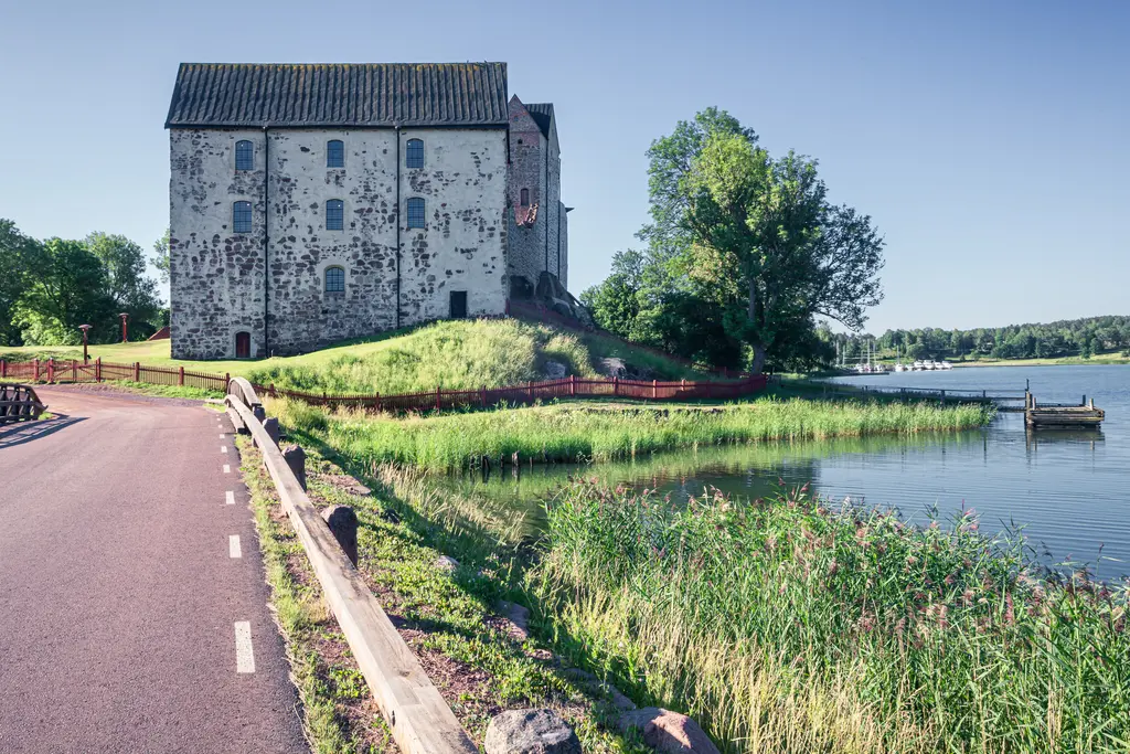 Historisches Gebäude am Wasser, umgeben von Grünflächen und Bäumen, mit einer ruhigen Straße im Vordergrund.