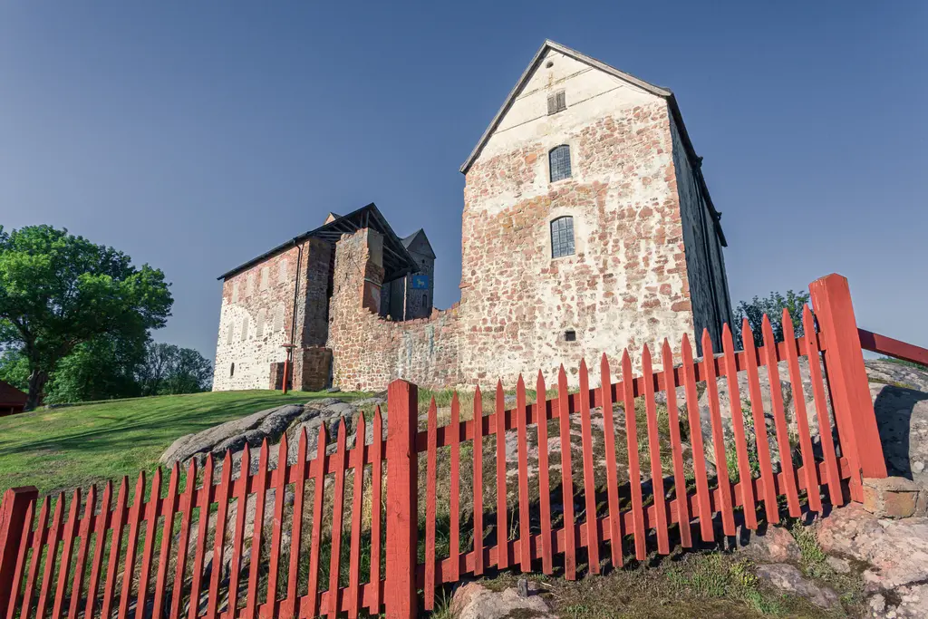 Rot eingezäunte Wiese mit einer historischen Steinruine, die teilweise erhalten ist. Im Hintergrund große Bäume unter klarem Himmel.