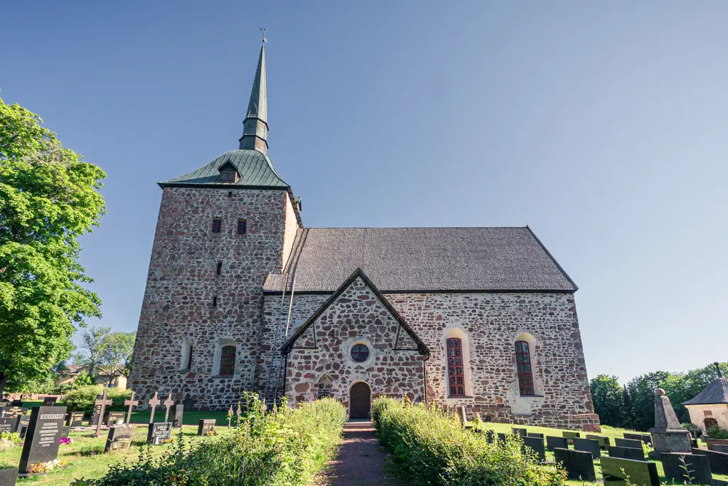Steinerne Kirche mit spitzem Turm und Satteldach, umgeben von einem gepflegten Garten und Grabsteinen. Klare, blaue Himmel.