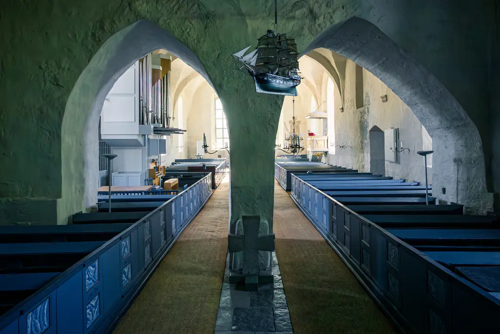 Innenansicht einer Kirche mit hohen Bögen und blau gestrichenen Bänken. Ein Modellschiff hängt von der Decke, und an den Wänden sind Kirchenorgeln sichtbar.