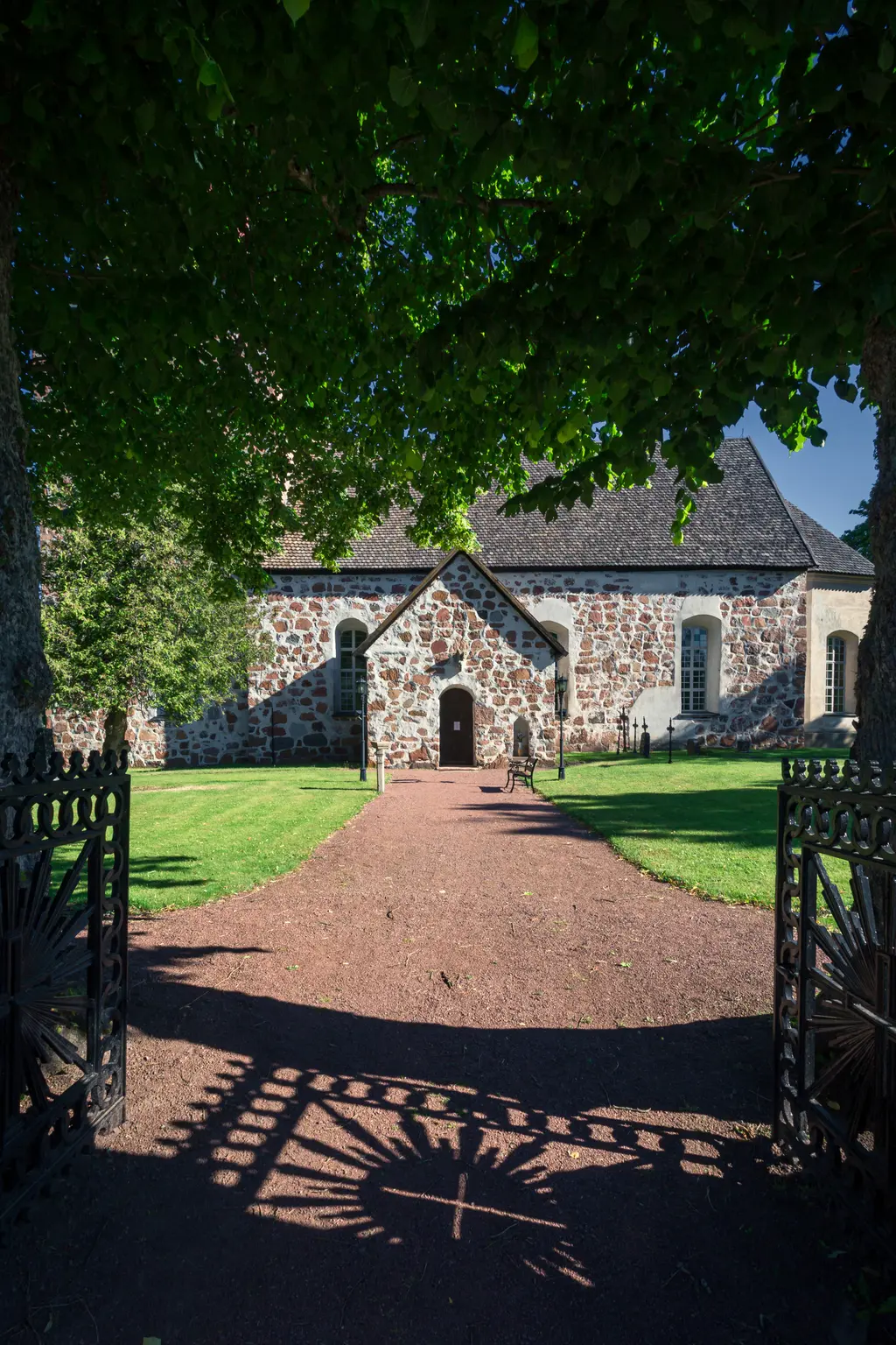 Historisches Gebäude mit steinernem Fassaden und spitzem Dach, umgeben von grünem Gras und Bäumen. Ein geschwungener Weg führt zum Eingang.