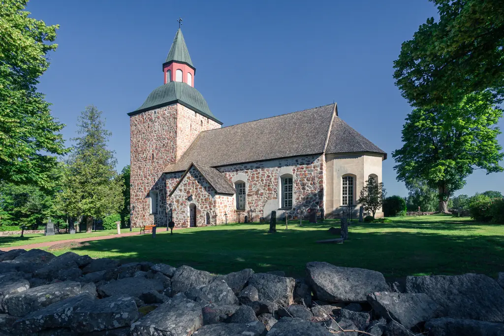 Steinige Kirche mit einem rotgedeckten Turm, umgeben von Bäumen und einem gepflegten Garten. Felsen im Vordergrund.