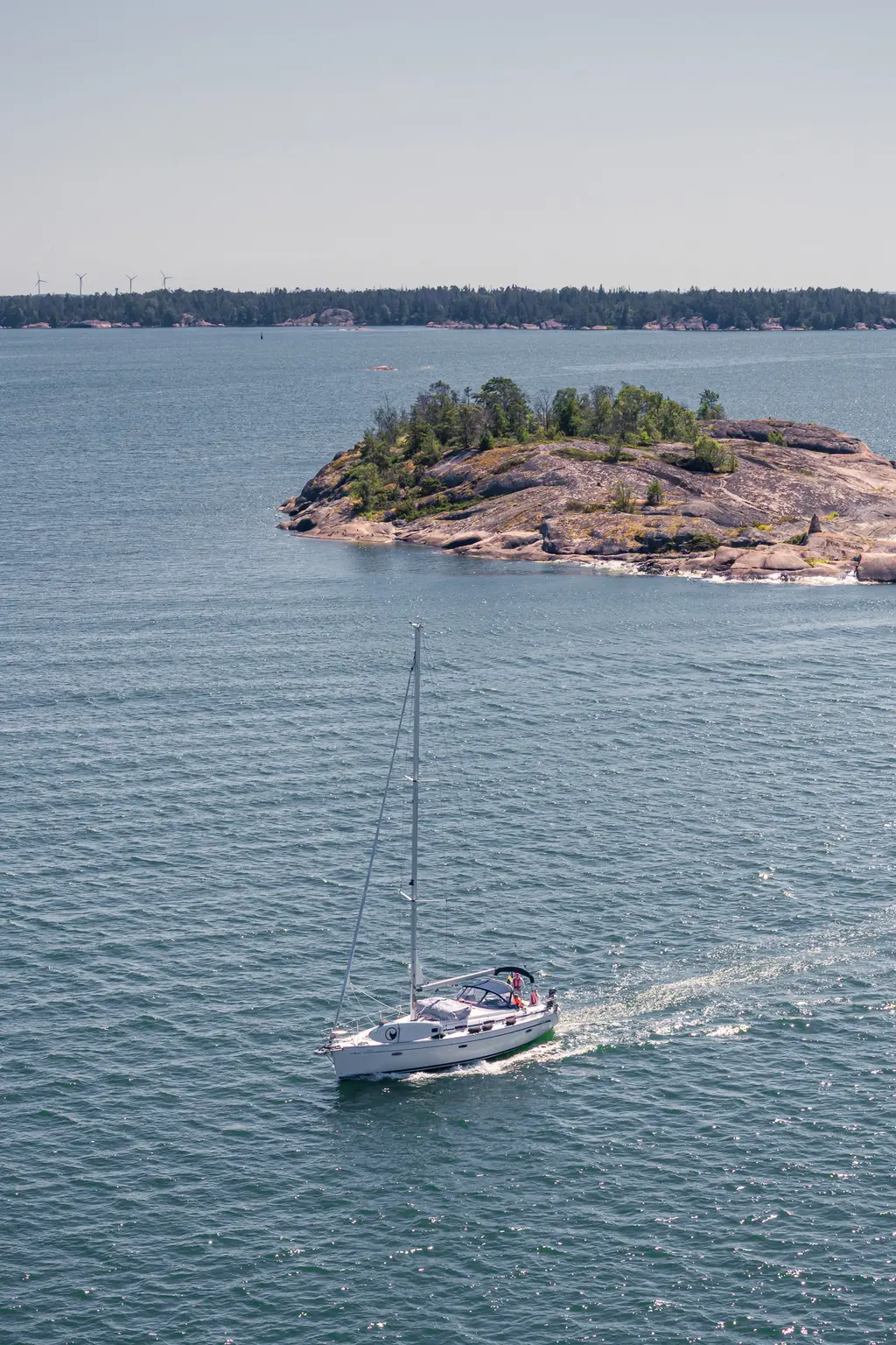 Segelboot auf ruhigem Wasser, vorbei an einer kleinen, bewaldeten Insel. Im Hintergrund sind sanfte Hügel und weitere Inseln sichtbar.