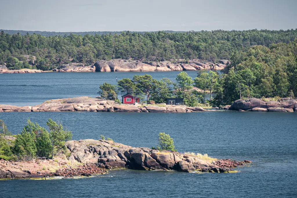 Rote Holzhütte auf einer grünen Insel, umgeben von blauem Wasser und Felsen, mit bewaldetem Ufer im Hintergrund.