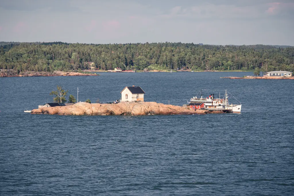 Insel mit einem kleinen, weißen Haus und einem Schiff, umgeben von Wasser und bewaldeten Ufern im Hintergrund.