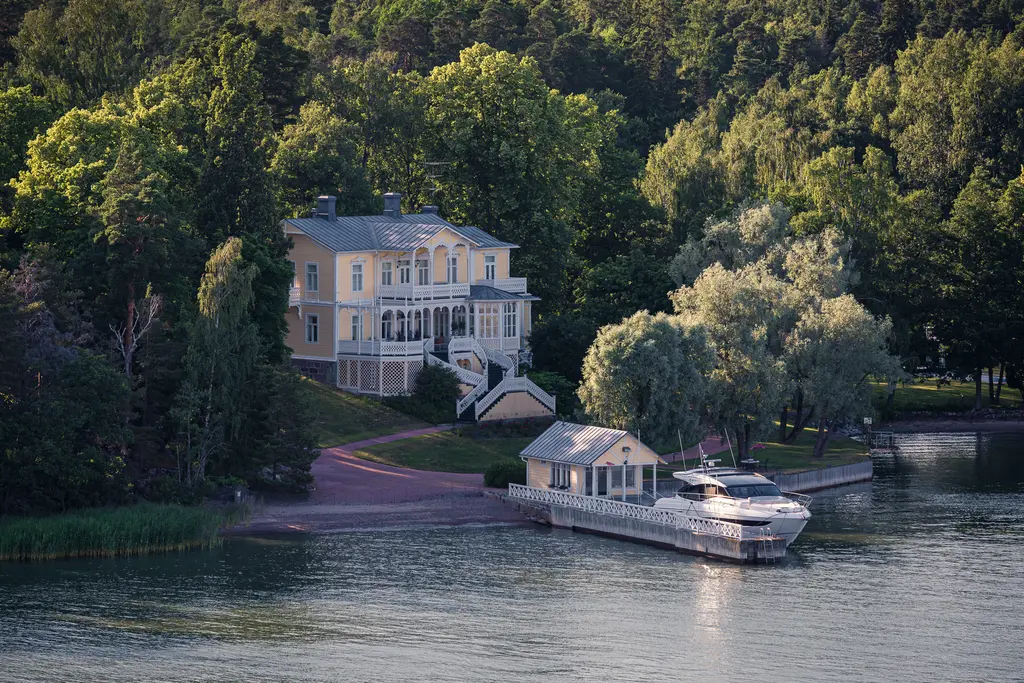 Helles, elegantes Haus mit Veranda steht am Ufer, umgeben von Bäumen. Am Wasser liegt ein kleiner Bootsanleger mit einem Boot.