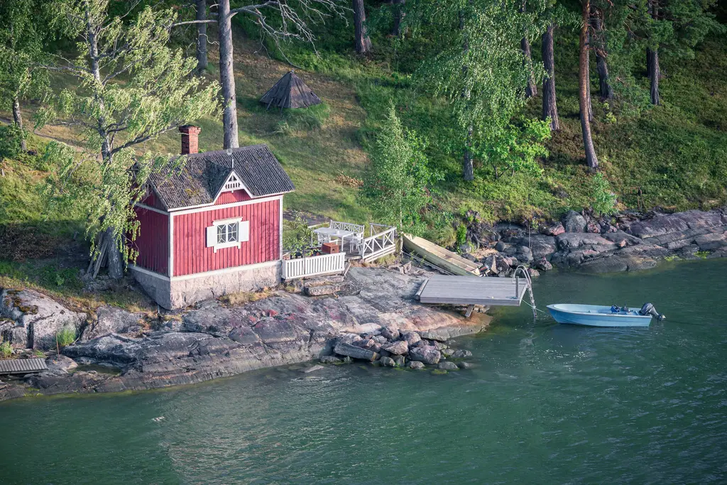 Rotes Sommerhaus am Wasser mit Steg und kleiner Motorboot in einer ruhigen, bewaldeten Landschaft. Grüne Bäume umgeben die Szenerie.