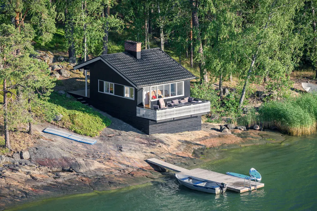 Ein schwarzes Holzhaus steht auf Felsen am Ufer, umgeben von Bäumen. Eine kleine Holzterrasse bietet Blick auf das Wasser, daneben liegt ein Boot.