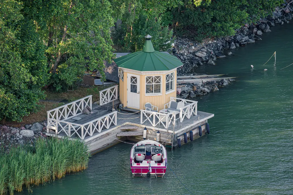 Kleine, rundliche Holzplattform am Wasser mit einem grünen Dach, umgeben von Bäumen und Steinen. Ein Boot liegt am Steg.