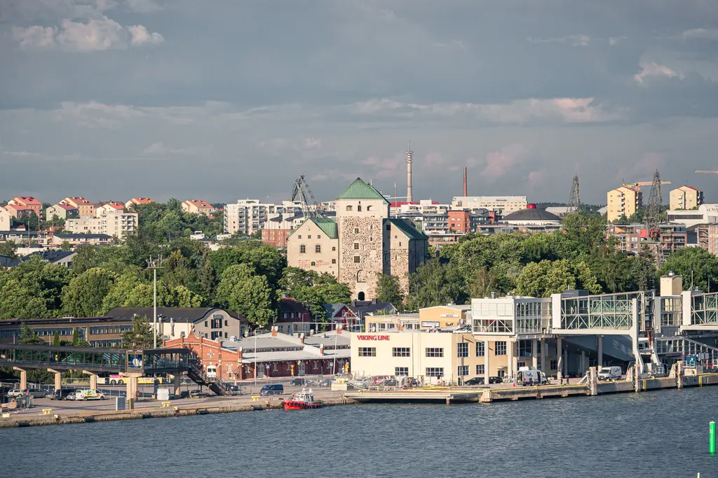Blick auf ein historisches Schloss, umgeben von Bäumen, mit modernen Gebäuden im Hintergrund und einem Hafenbereich im Vordergrund.