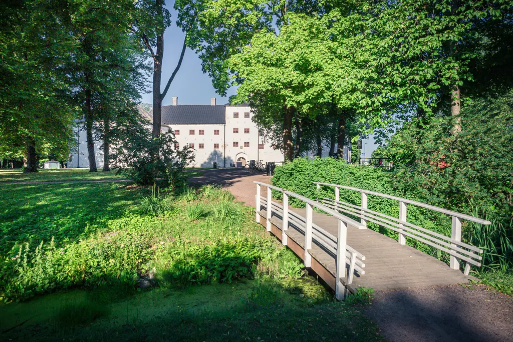 Brücke über einen kleinen Wasserlauf, umgeben von grünem Gras und Bäumen. Im Hintergrund ein historisches Gebäude mit weißen Wänden.