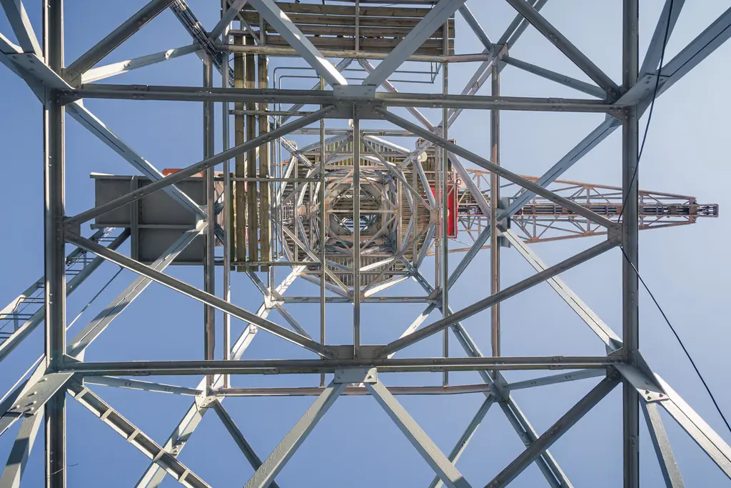 Blick nach oben durch die Struktur eines Metallmastes, der von zahlreichen Stahlträgern umgeben ist, mit klarem Himmel im Hintergrund.