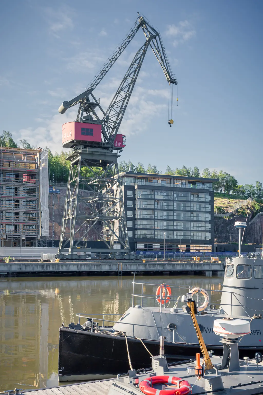 Hafenkran neben einem modernen Gebäude und einem schwarzen Boot im Wasser. Im Hintergrund sind Baustellen und Bäume zu sehen.