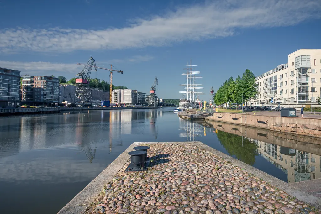 Hafenansicht mit geschäftigen Kränen und modernen Gebäuden. Ein historisches Segelschiff liegt ruhig im Wasser, umgeben von Bäumen.
