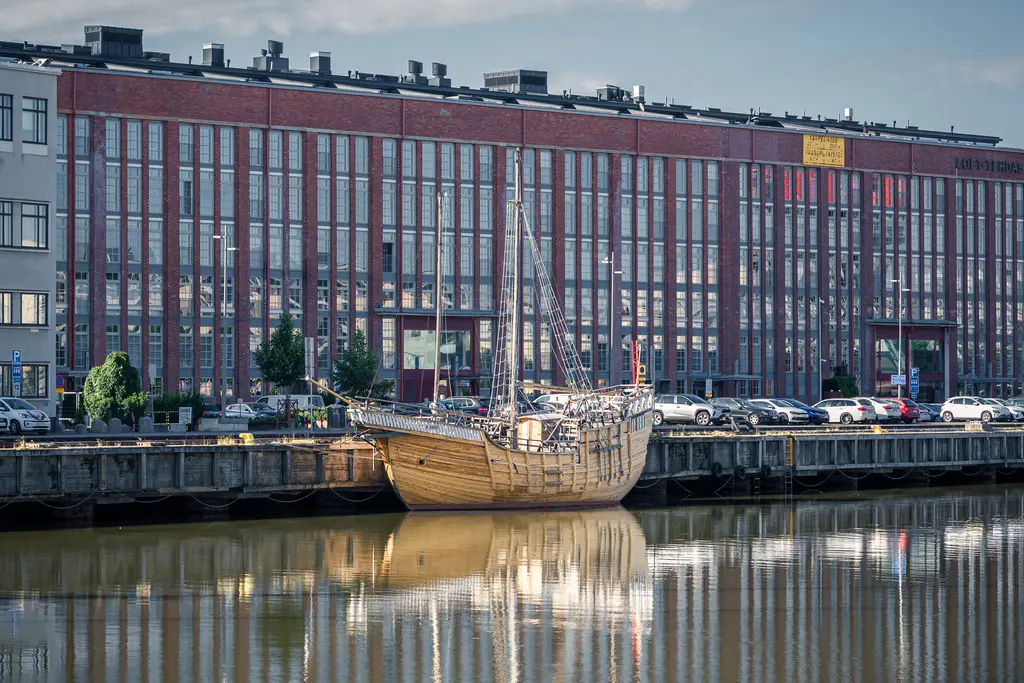 Historisches Segelschiff am Ufer eines ruhigen Gewässers, gegenüber einer modernen Backsteinarchitektur mit Fensterfronten.