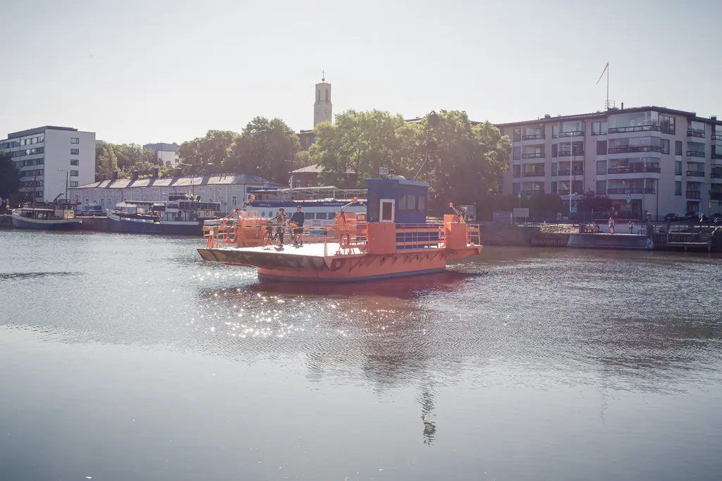 Fähre fährt über das Wasser, umgeben von Bäumen und modernen Gebäuden. Die Oberfläche des Wassers glänzt in der Sonne.