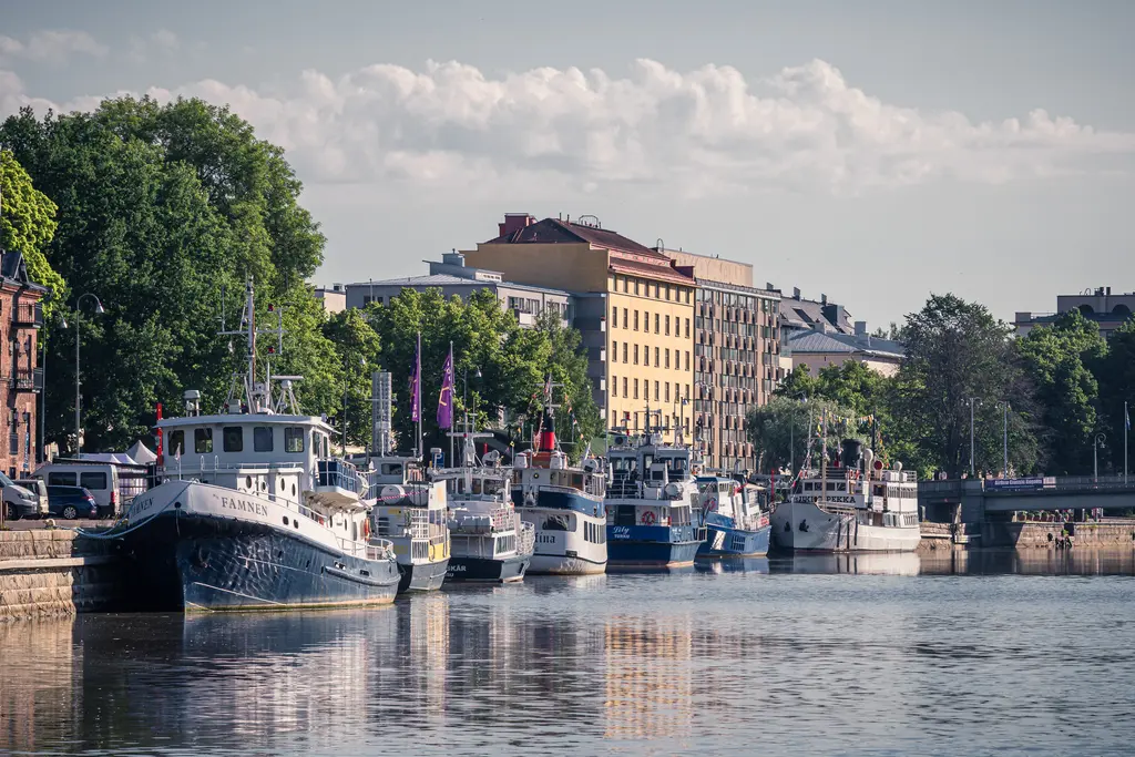 Boote liegen am pier, umgeben von grünen Bäumen und modernen Gebäuden. Ruhige Wasser spiegeln das Ufer wider.