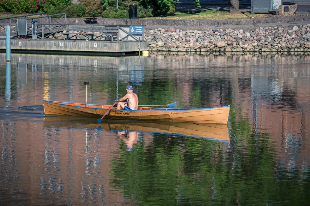 Ein Ruderer in einem hölzernen Boot paddelt ruhig auf einem klaren Gewässer, das die Umgebung reflektiert. Die Ufer sind von Bäumen und Steinen gesäumt.