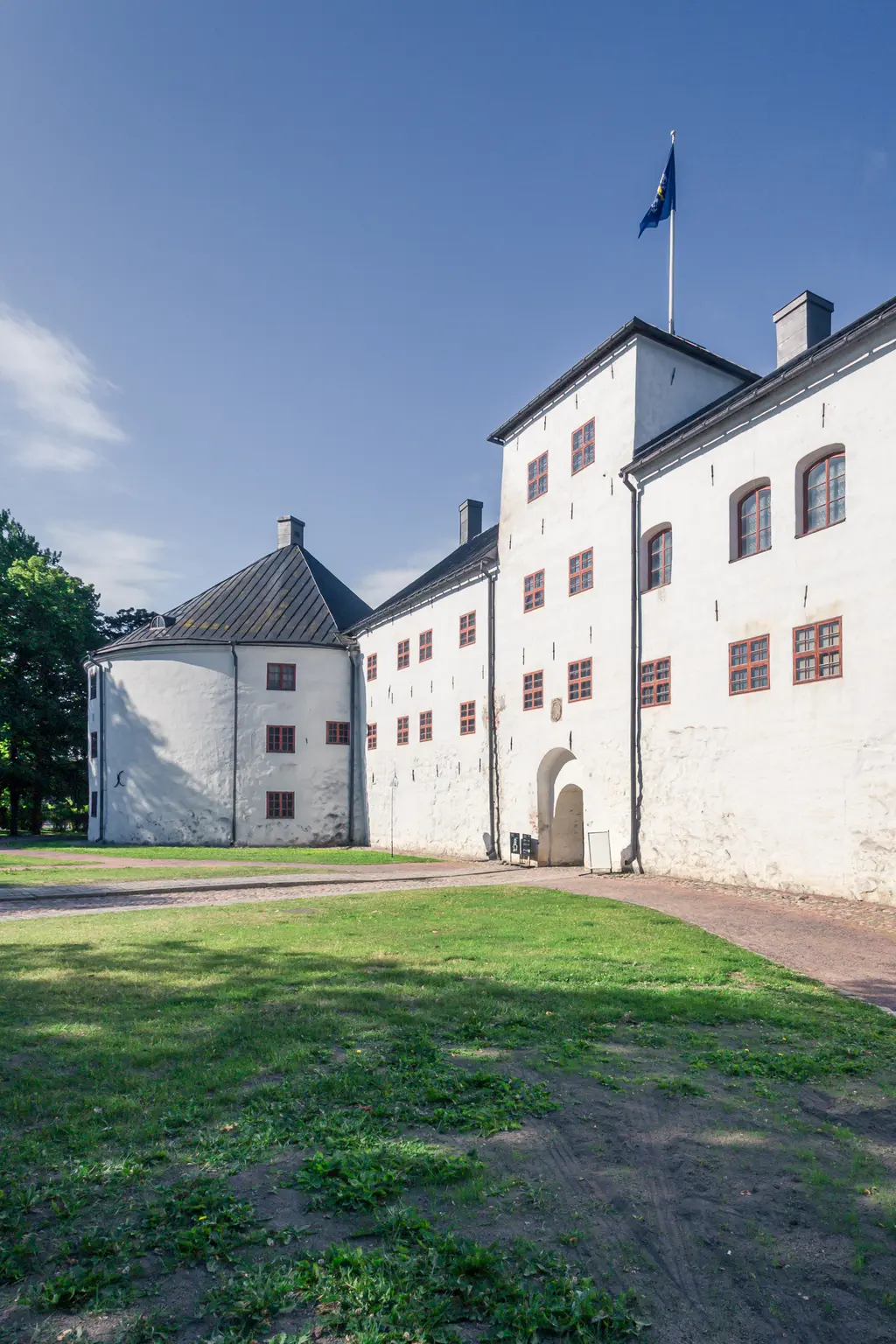 Historisches Gebäude mit weißer Fassade, roten Fenstern und einer Fahne, umgeben von einem grünen Rasen und einem Kiesweg.