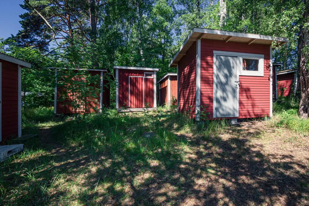 Rote Holzhütten umgeben von grünem Gras und Bäumen. Einige Hütten haben Fenster, eine ist mit einer grauen Tür ausgestattet.