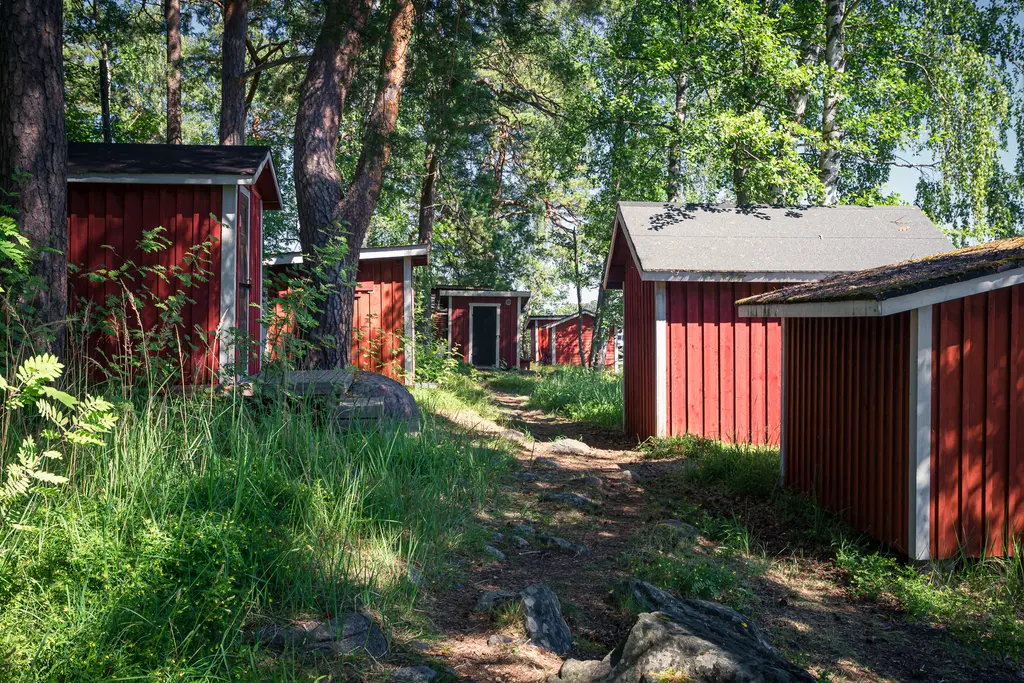 Rote Holzhäuser umgeben von grüner Vegetation und Bäumen, entlang eines schmalen Pfades, der durch die Anlage führt.