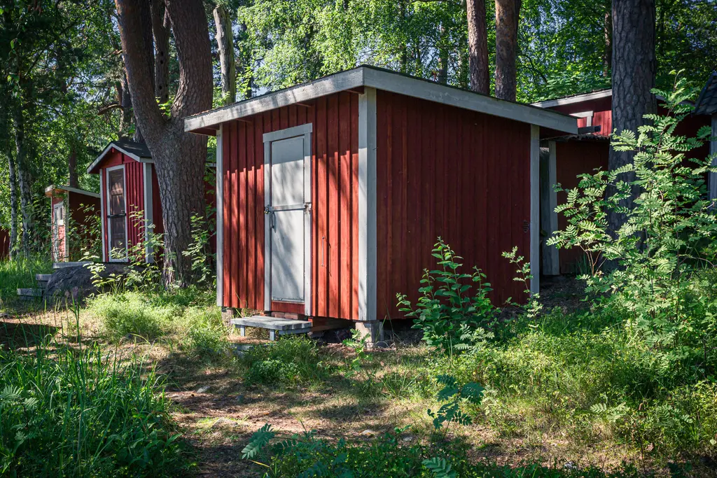 Rote Holzhütte im Wald, umgeben von Bäumen und Unterholz. Eine weitere Hütte ist im Hintergrund sichtbar.