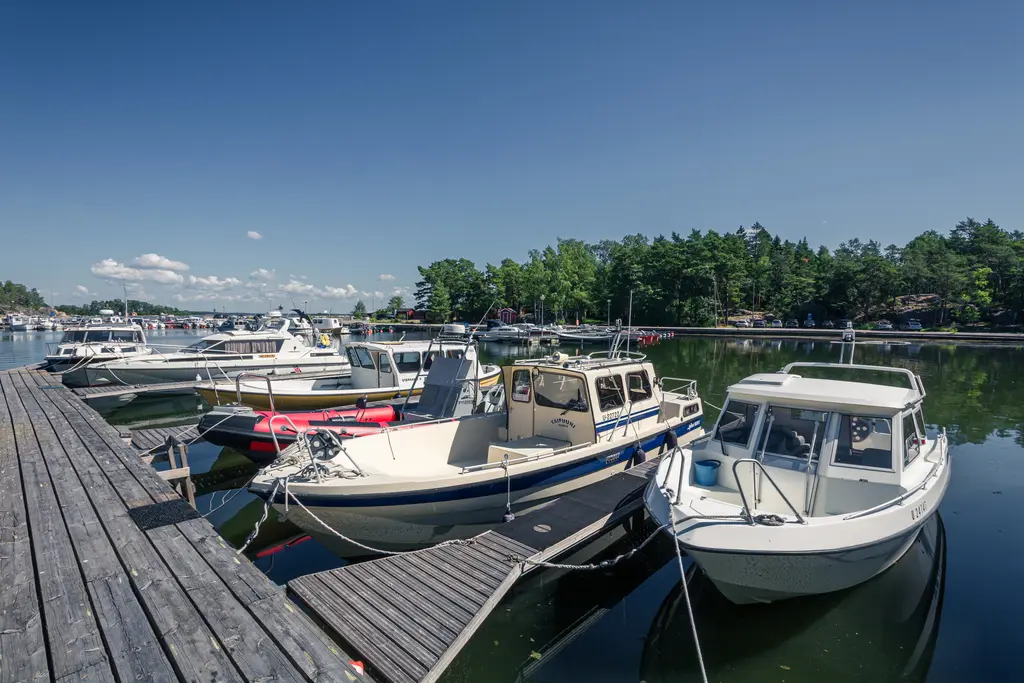 Boote im Hafen vor einem Holzsteg, umgeben von Bäumen und klarem Wasser. Ein blauer Himmel mit wenigen Wolken ist sichtbar.