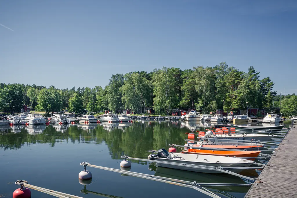 Hafen mit mehreren Booten, die an einem Steg festgemacht sind. Umgeben von Bäumen und spiegelndem Wasser unter klarem Himmel.