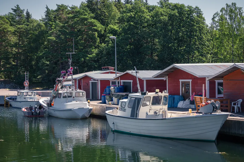 Fischerboote liegen im ruhigen Wasser an einem roten Holzsteg, umgeben von grünen Bäumen. Im Hintergrund stehen einfache, alte Häuser.
