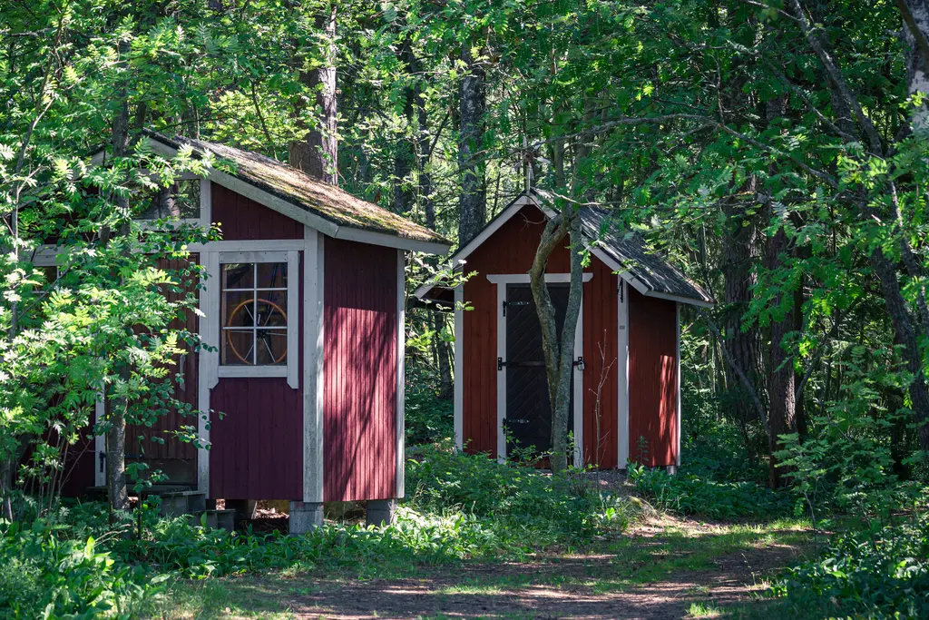 Zwei rote Holzhütten stehen umgeben von üppigem Grün und Bäumen. Sonnenlicht fällt auf die strahlenden Wände und den Boden.