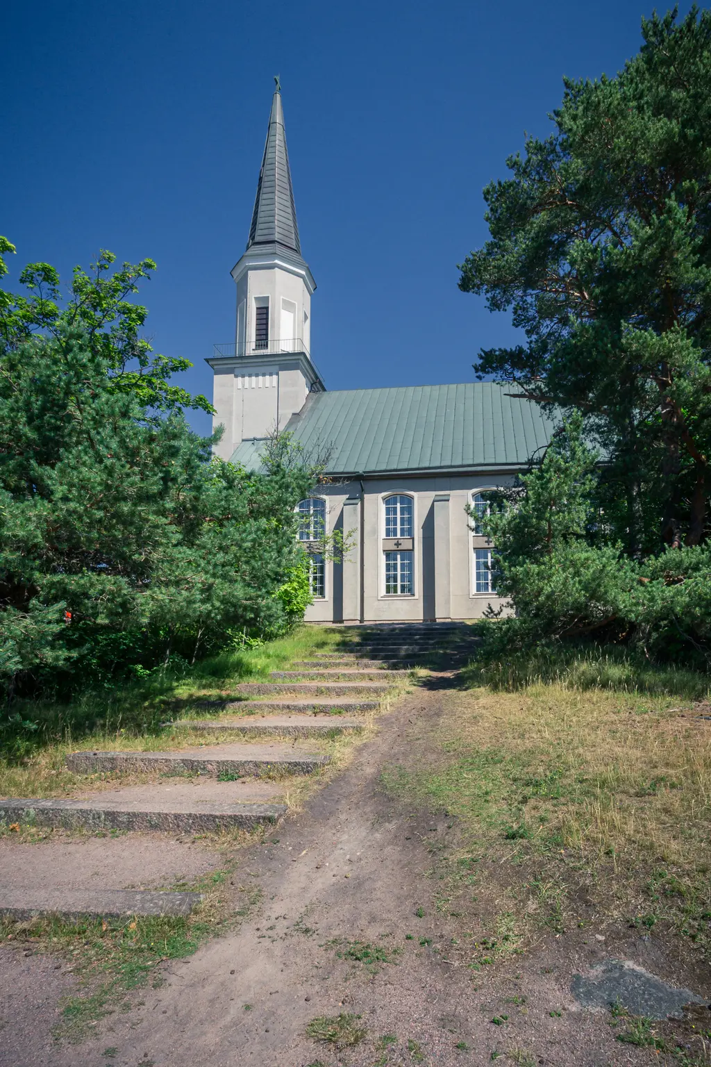 Kirche mit spitzem Glockenturm, von Stufen umgeben. Grüne Bäume und Rasen säumen den Weg zur Eingangstür.