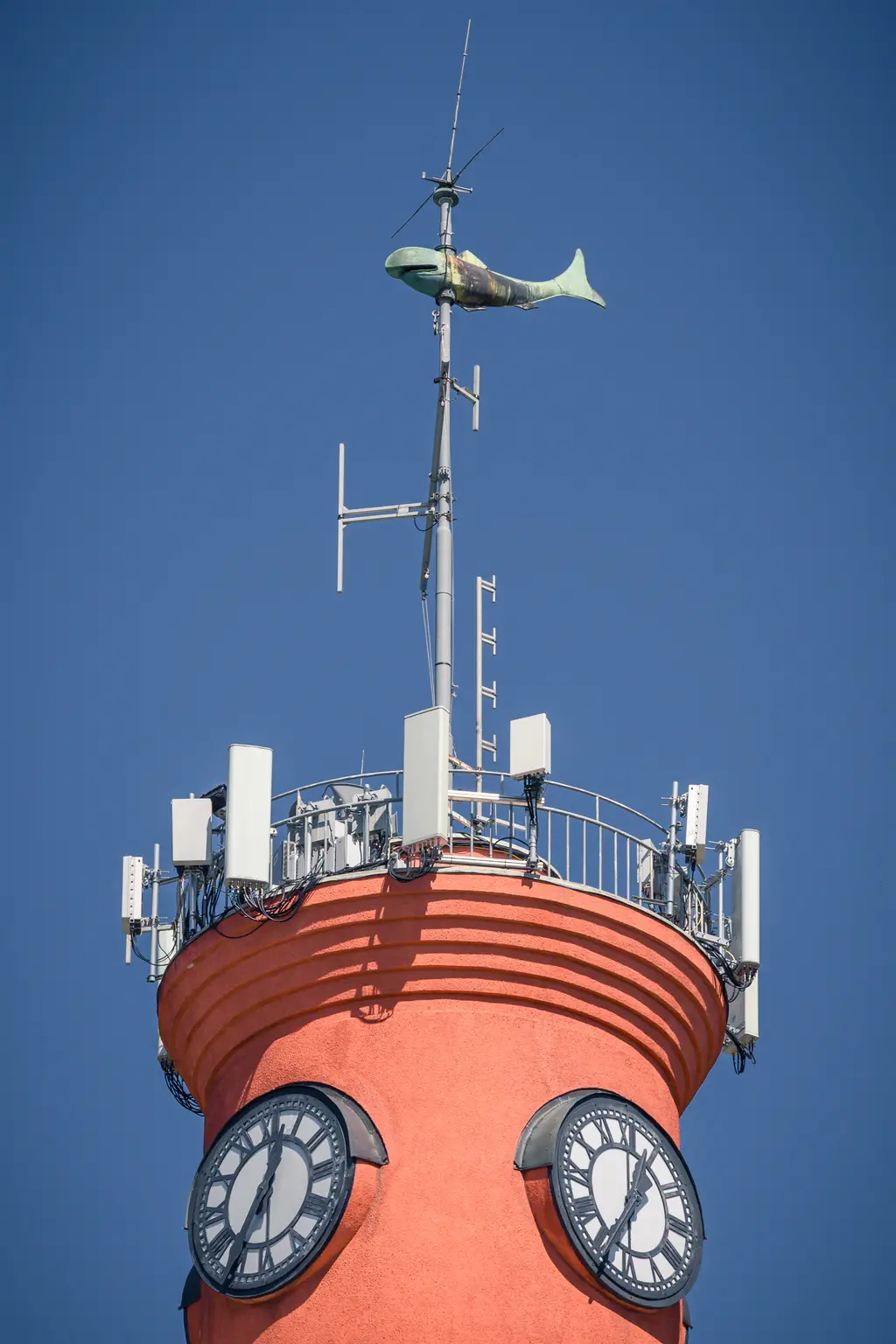 Mast mit leuchtend roter Färbung, ausgestattet mit mehreren Antennen und zwei großen Ziffernblättern. Oben sitzt eine Fischskulptur.