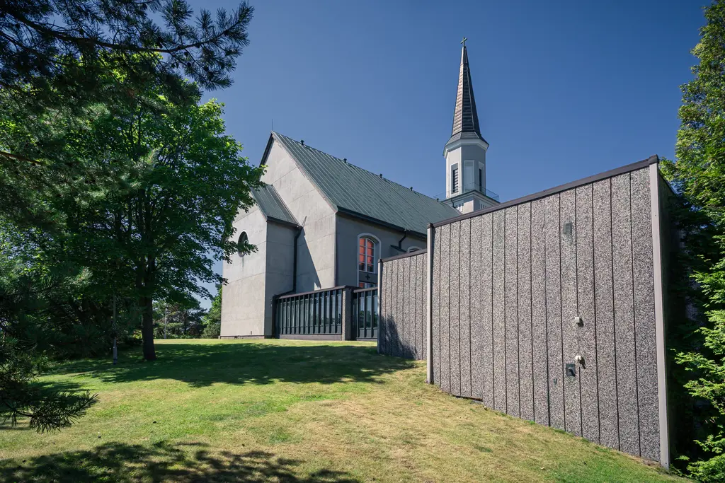 Kirche mit einem hohen Turm, umgeben von Bäumen und einer Wiese. Neben der Kirche steht eine moderne, graue Wand aus Holz.