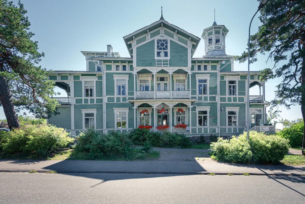 Holzhaus im Jugendstil mit grüner Fassade, verzierenden Balkonen und Blumenbalkonen. Das Gebäude hat viele Fenster und einen kleinen Turm.