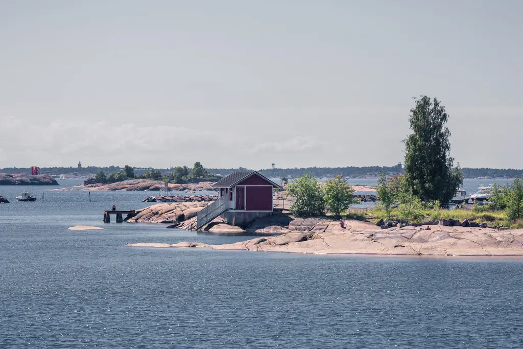 Rotes Holzhaus mit Treppe auf einer kleinen Felseninsel, umgeben von Wasser und etwas Vegetation, im Hintergrund weitere Inseln.