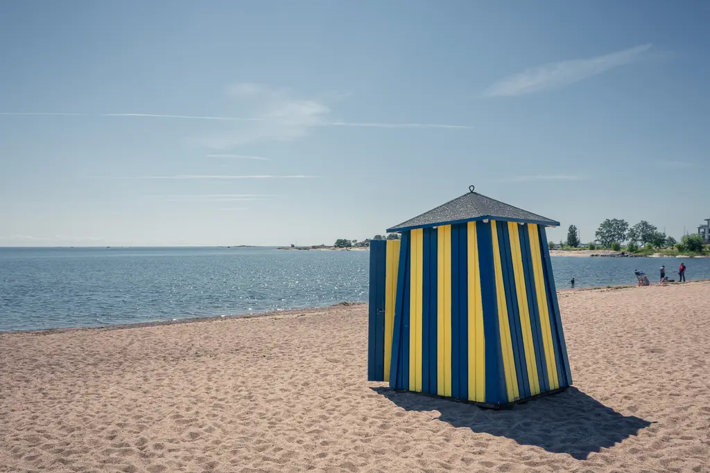 Strandhäuschen mit blauen und gelben Streifen steht auf einem Sandstrand. Im Hintergrund erstreckt sich das glitzernde Wasser.