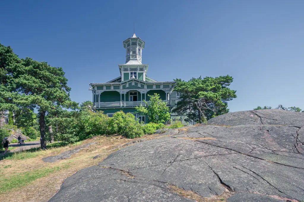 Historisches Gebäude mit grünem Holzverblendung, Türmchen und großen Fenstern, umgeben von Bäumen und Felsen.