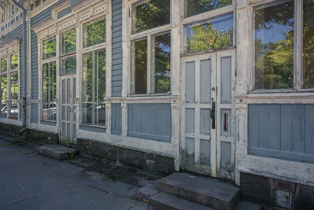 Fassade eines alten, blauen Holzgebäudes mit großen Fenstern und weißen Türen. Der Boden ist mit Steinen gepflastert.